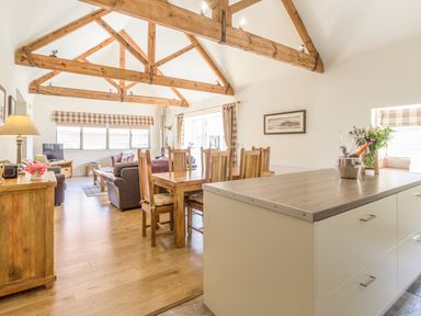 An open plan living and dining area with exposed wooden beams a dining table chairs sofas and a kitchen island at Sawmill Cottage in Belford near Bamburgh