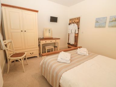 A bedroom with a bed covered in a striped blanket and towels a wardrobe a chair a dressing table with a stool a wall-mounted TV and robes hanging on a wooden door at Sawmill Cottage in Belford near Bamburgh