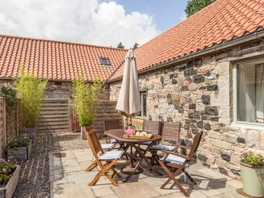 An outdoor patio with a round wooden table and six wooden chairs with cushions next to a stone building at Sawmill Cottage in Belford near Bamburgh
