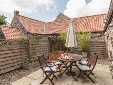 An outdoor patio area with wooden chairs and a table with an umbrella and food at Sawmill Cottage in Belford near Bamburgh
