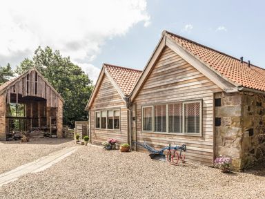 A wooden house with red-tiled roof and gravel ground with plants and an old farming tool at Sawmill Cottage in Belford near Bamburgh