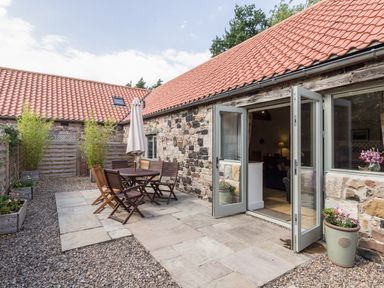 A stone patio with wooden table and chairs and umbrella outside a house with tiled roof at Sawmill Cottage in Belford near Bamburgh