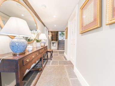 A hallway with a wooden table holding lamps and plants a round mirror on the wall stone floor and framed pictures at Prospect House in Ambleside