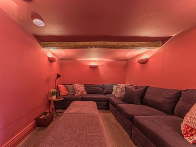 A small seating room with a large dark sectional sofa with cushions a small round side table with a flower pot and a lamp and wall lights and wooden ceiling beam at Prospect House Ambleside