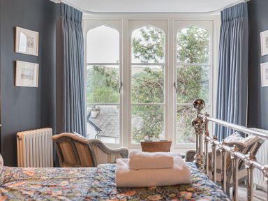 A bedroom with a metal bed frame two armchairs a wooden chest large windows with curtains and views of trees at Prospect House in Ambleside
