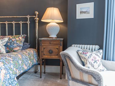 A bedroom with a metal bed frame floral bedding a wooden bedside table with a lamp a chair with cushions and blue curtains at Prospect House Ambleside