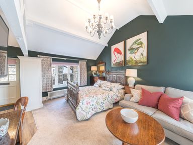 A bedroom with a wooden bed, floral bedding, a beige sofa with red and beige pillows, wooden tables, and bird paintings on a green wall at Prospect Cottage Loft in Ambleside