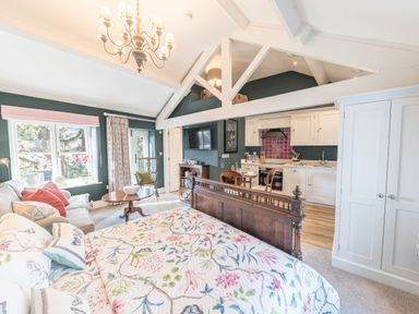 A bedroom with a wooden bed frame and floral bedding next to a sofa a round wooden table and a rocking chair with a kitchen in the background at Prospect Cottage Loft in Ambleside