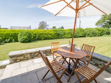 A patio with a wooden table and chairs under a white umbrella with a grassy lawn and shrubs in the background at Berwyn in Talybont