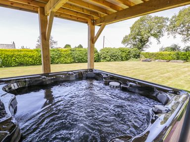 A hot tub under a wooden pergola overlooking a grassy backyard with hedges and trees at Berwyn in Talybont