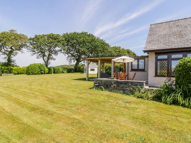 An outdoor patio with wooden chairs and a table under an umbrella next to a house and a large lawn with trees at Berwyn in Talybont