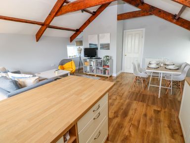 An open living and dining room with wooden beams on the ceiling a wooden floor a gray sofa a yellow chair a television on a white shelf and a dining table with white chairs at The Old Brewery in Saundersfoot