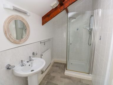 A bathroom with a shower, a sink with two taps, a round mirror, a radiator, and a wooden ceiling beam at The Old Brewery in Saundersfoot