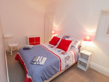 A bedroom with a double bed with patterned bedding red pillows bedside tables with lamps a white chair and a storage unit at 1 Church Square in Whitby