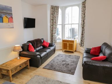 A living room with two black sofas with red cushions a wooden coffee table a small wooden cabinet under a large window and a mounted flat screen TV at 1 Church Square Whitby