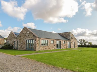 A stone building with multiple windows and doors surrounded by grass and a gravel path at The Long Barn in Berwick-Upon-Tweed