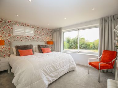 A bedroom with a large bed that has white bedding and orange pillows and two matching orange lamps and an orange chair by a large window at Shutts House Garden Apartment in Coombe Bissett