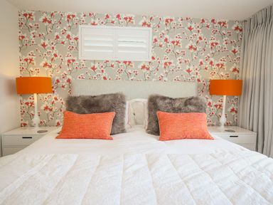 A bedroom with a bed featuring white bedding orange and gray pillows floral wallpaper and two orange lamps at Shutts House Garden Apartment in Coombe Bissett