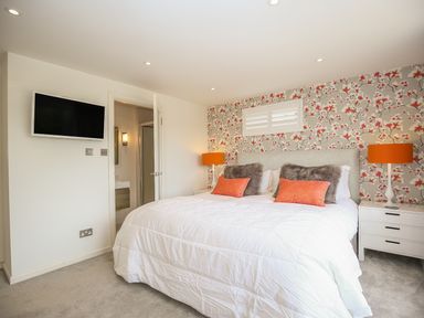 A bedroom with a bed white bedding floral wallpaper orange lamps and a wall-mounted tv at Shutts House Garden Apartment in Coombe Bissett