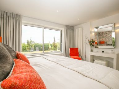 A bedroom with a bed, orange cushions, an orange chair, a dressing table with a mirror and a window with curtains at Shutts House Garden Apartment in Coombe Bissett