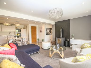 A living room with a blue sofa, glass coffee table, white armchairs, and a kitchen with a dining table in the background at Shutts House Garden Apartment in Coombe Bissett
