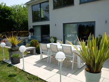 An outdoor patio with a white dining table and chairs surrounded by plants and a modern house with large windows in the background at Shutts House Garden Apartment in Coombe Bissett