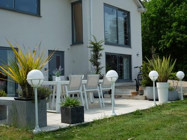 An outdoor patio with white chairs and table surrounded by potted plants and globe lights at Shutts House Garden Apartment in Coombe Bissett
