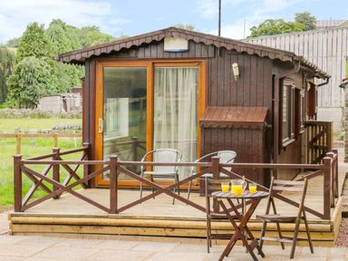 A wooden cabin with a deck and sliding glass door with two chairs on the deck and a small table with two chairs and a jug of orange juice in front at Thirley Beck Lodge in Harwood Dale