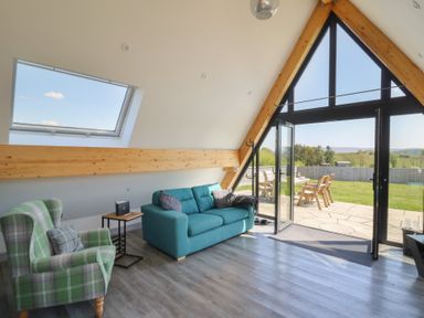A living room with a green plaid armchair a blue sofa and large triangular windows opening to a patio at Elan Valley Welsh - The Sheepfold in Llanyre near Llandrindod Wells