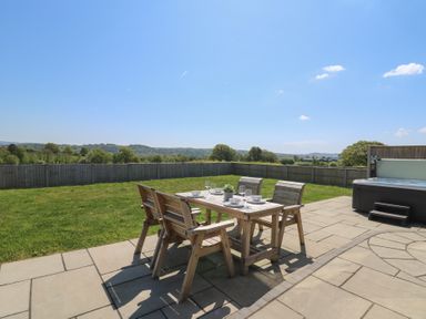 A patio with a wooden dining table and chairs on a lawn with a hot tub at Elan Valley Welsh - The Sheepfold in Llanyre near Llandrindod Wells