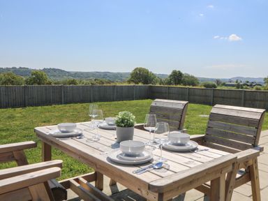An outdoor wooden dining table set with plates bowls cutlery wine glasses and a plant with a fenced grass yard and hills in the background at Elan Valley Welsh - The Sheepfold in Llanyre near Llandrindod Wells