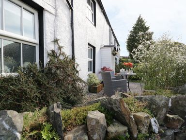An outdoor seating area with a small table and chair beside a white house with plants and a stone wall at Rusland View Rusland near Newby Bridge