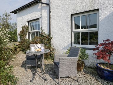 An outdoor seating area with a round glass table and two wicker chairs beside a white house with two windows at Rusland View Rusland near Newby Bridge
