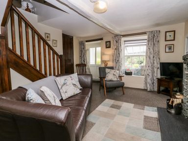 A living room with a leather sofa and armchair beside a wooden staircase and window with curtains at Rusland View Rusland near Newby Bridge