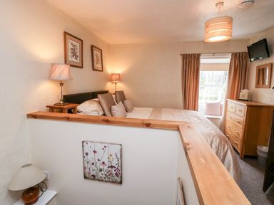 A bedroom with a bed, lamps, framed pictures, a dresser, window with curtains and a wall-mounted TV at Rusland View Rusland near Newby Bridge