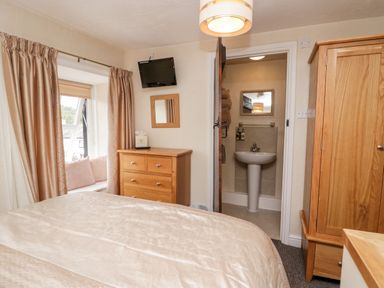 A bedroom with a bed a wooden dresser a wall-mounted TV window with curtains and a view and a doorway leading to a bathroom at Rusland View Rusland near Newby Bridge