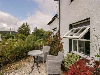 An outdoor seating area with a round glass table and two wicker chairs beside a white building with open windows at Rusland View Rusland near Newby Bridge