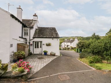 A white cottage with potted plants outside near a paved road with more white houses and green trees in the background at Rusland View Rusland near Newby Bridge