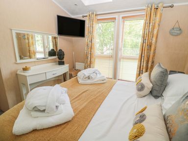 A bedroom with a bed covered in white and beige linens and pillows with floral decorations a dresser with a mirror and a Buddha head sculpture a wall-mounted television and windows with yellow floral curtains at Ramblers Rest in Felton