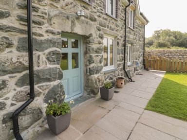 A stone exterior wall with a light blue door and windows with flower pots on a stone patio at Tyn Ffynnon in Dyffryn Ardudwy