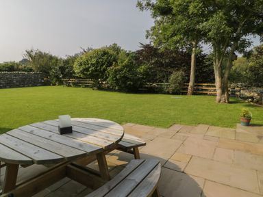 A garden with a wooden picnic table on a stone patio and grass lawn with trees and bushes at Tyn Ffynnon in Dyffryn Ardudwy