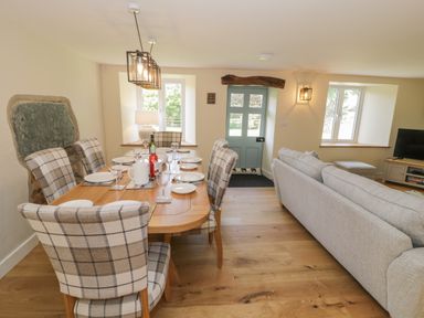 A dining area with a wooden table set with plates and glasses and plaid chairs next to a living area with a gray sofa and a television at Tyn Ffynnon in Dyffryn Ardudwy