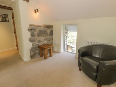 A room with a black armchair a wooden stool a stone wall section and a window at Tyn Ffynnon in Dyffryn Ardudwy