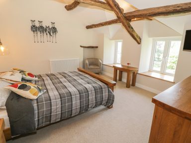 A bedroom with a wooden bed, patterned pillows, a gray chair, wooden table, and two windows at Tyn Ffynnon in Dyffryn Ardudwy