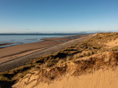 A sandy beach with dunes and grass by a calm sea with mountains in the distance at Tyn Ffynnon in Dyffryn Ardudwy