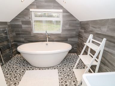 A bathroom with a freestanding bathtub under a window with blinds and a white shelf unit on patterned floor tiles at Argraig in Llanbedrog