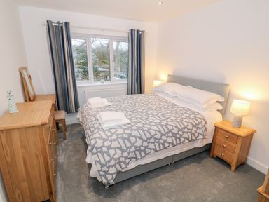 A bedroom with a bed covered with a patterned blanket two bedside tables with lamps a wooden dresser and a window with curtains at Argraig in Llanbedrog