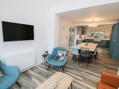 An open plan living room and kitchen with blue chairs a wooden dining table a wall mounted tv and wood flooring at Argraig in Llanbedrog