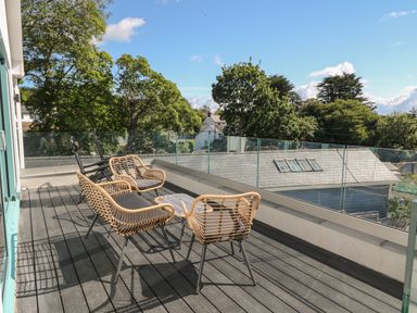 An outdoor balcony with wicker chairs and a table overlooking trees and rooftops at Argraig in Llanbedrog