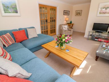 A living room with a blue sectional sofa a wooden coffee table with flowers a TV on a glass stand and a striped armchair at Henrys Croft in Lizard